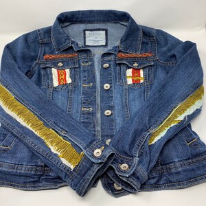Peut inclure: Une veste en jean bleue avec des bordures dor&eacute;es et blanches sur les manches et des bordures rouges, blanches et dor&eacute;es sur les poches. La veste porte une &eacute;tiquette indiquant "Sonoma Life + Style" et "XS".