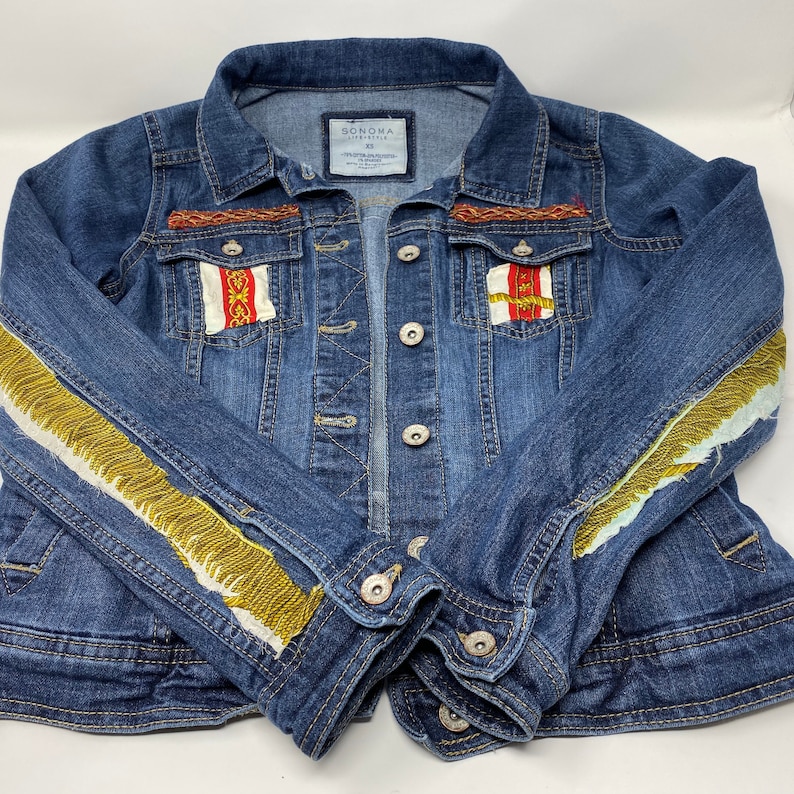 Peut inclure: Une veste en jean bleue avec des bordures dor&eacute;es et blanches sur les manches et des bordures rouges, blanches et dor&eacute;es sur les poches. La veste porte une &eacute;tiquette indiquant "Sonoma Life + Style" et "XS".
