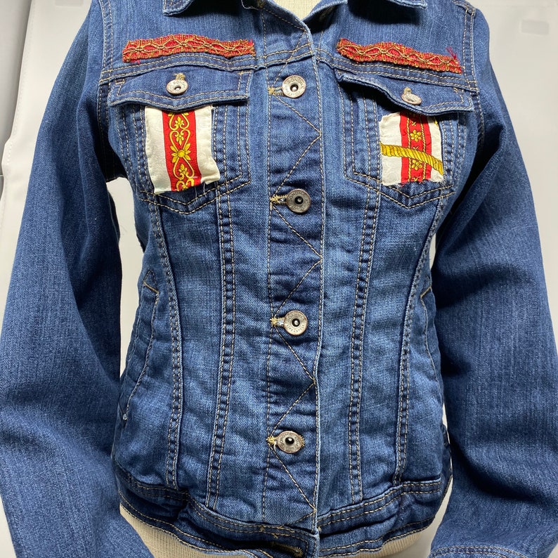 Peut inclure: Une veste en jean bleue avec des bordures rouges et dor&eacute;es sur les poches et le col. La veste a une fermeture &agrave; boutons et des manches longues.