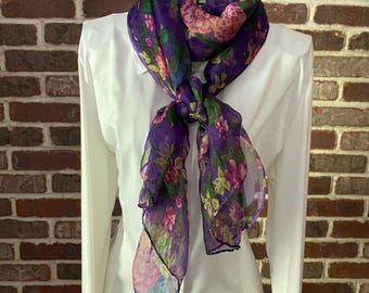 May include: A sheer purple scarf with a floral pattern of pink, green, and blue flowers. The scarf is draped over a white collared shirt and tied in a knot. The background is a brick wall.