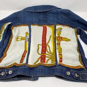 Peut inclure: Une veste en jean bleue avec un tissu &agrave; motifs blanc, rouge et or cousu sur le dos. Le tissu pr&eacute;sente un motif r&eacute;p&eacute;t&eacute; d'une corde, d'une &eacute;p&eacute;e et d'un b&acirc;ton d&eacute;coratif.