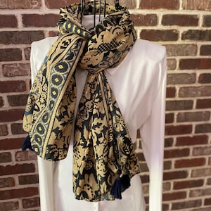 May include: A long scarf featuring a repeating pattern of black and gold floral and geometric motifs. The scarf is draped around the neck and includes dark blue tassels at the ends. It is displayed on a white collared shirt against a brick wall.