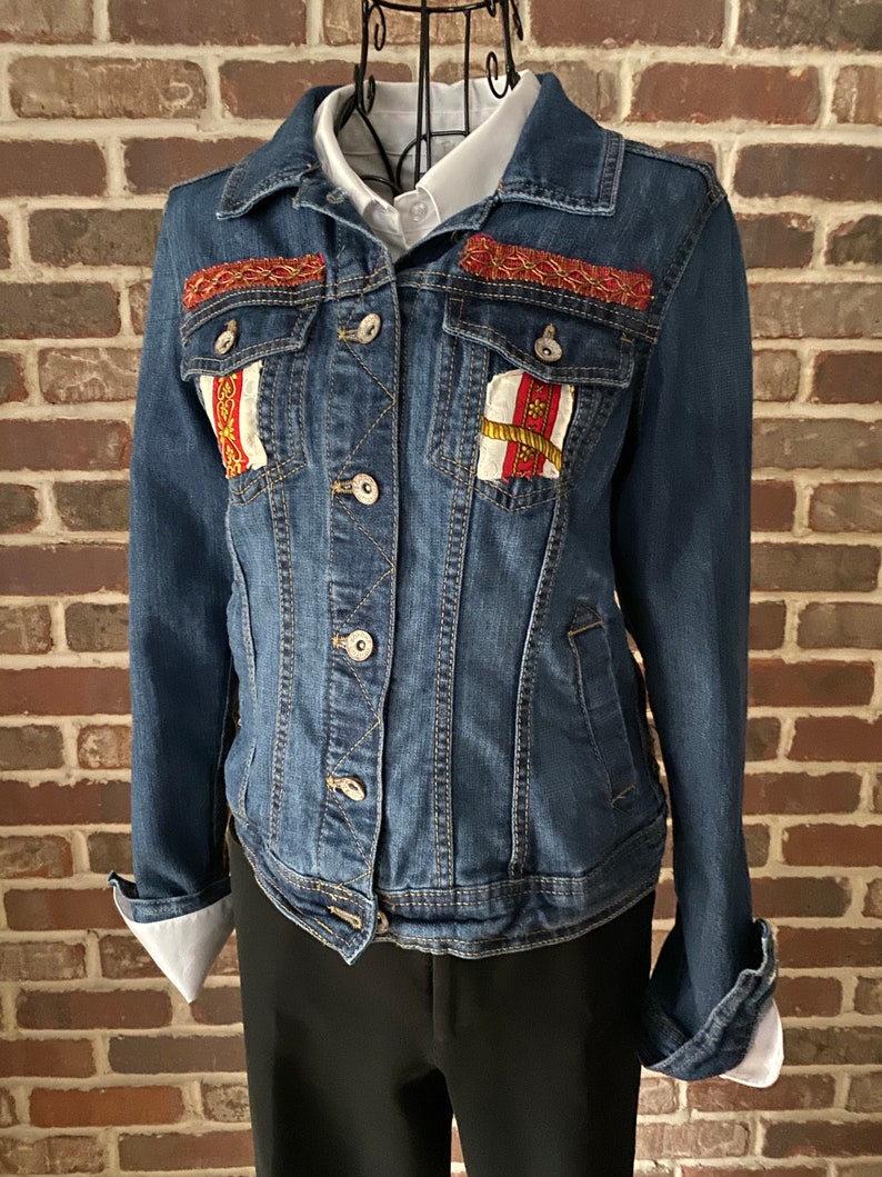 Peut inclure: Une veste en jean bleue avec des d&eacute;tails brod&eacute;s rouges, blancs et dor&eacute;s sur les poches avant et le col.