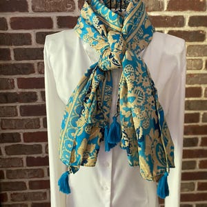 May include: A bright turquoise and beige scarf featuring a repeating floral and paisley pattern. The scarf is tied in a knot at the neck and finished with small turquoise tassels at the ends, draped over a plain white long-sleeved button-down shirt.