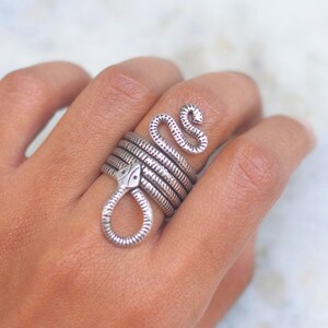 May include: Silver-toned snake ring. The ring wraps around the finger with a coiled design and a snake head at the top and bottom. The ring is worn on a hand with a light skin tone. The background is a blurred white and gray.