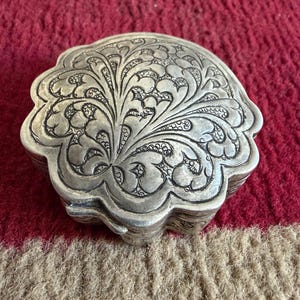 May include: A silver-colored, ornate box with a floral design. The box has a scalloped edge and is resting on a red and tan patterned surface. The box's surface is covered in detailed, engraved patterns.