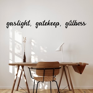 Peut inclure: Un bureau en bois avec une chaise en métal noir et une lampe blanche. Le mur derrière le bureau a le texte "gaslight, gatekeep, girlboss" écrit en cursive noire.