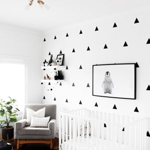 May include: A modern nursery with a white crib, gray armchair, and black and white patterned rug. The white walls are decorated with black triangle decals. A framed penguin print hangs above the crib.