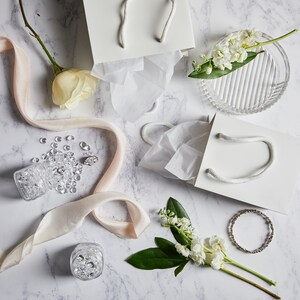 May include: White gift bags with tissue paper, a white ribbon, a small glass jar filled with clear rhinestones, a ring, and white flowers on a marble surface.
