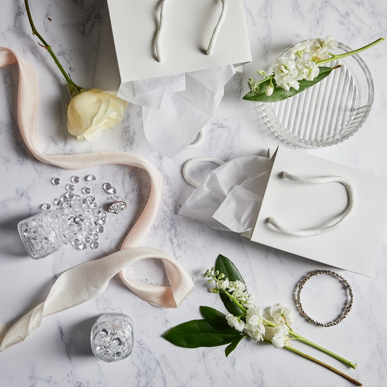May include: White gift bags with tissue paper, a white ribbon, a small glass jar filled with clear rhinestones, a ring, and white flowers on a marble surface.