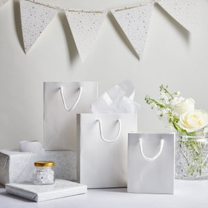 May include: Three white gift bags of different sizes with white handles. The bags are on a white surface with a small glass jar filled with white beads and a white gift box.