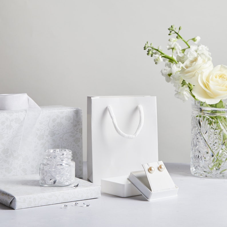 May include: White gift bag with white handles, a small white box with pearl earrings, a clear glass jar filled with clear crystals, and a white gift wrap with a silver pattern. A vase with white flowers is in the background.