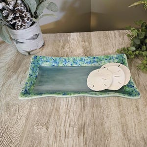 May include: A rectangular, sea-themed glass tray with a light blue center and a speckled green and blue border. Three white sand dollars rest on the tray. The tray is displayed on a wooden surface.