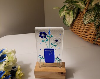Fused glass art pot of forever flowers in a lite stained wood base.  Made on clear glass with a blue pot of flowers and a white daisy.