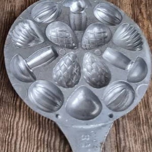 May include: A silver metal mold with a wooden handle. The mold has 12 individual cavities shaped like different nuts and mushrooms. The mold is open and ready to be used.