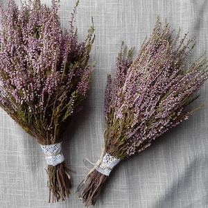 May include: Two bunches of dried heather flowers. The heather has small, light purple blooms and green stems. Each bunch is tied with twine and a white lace ribbon. The background is a light grey fabric.