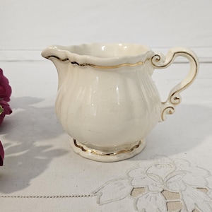 Puede incluir: Una jarrita para crema de cerámica color crema con borde festoneado, asa curva y cuerpo redondeado con acanaladuras verticales. El borde, la base y el asa están decorados con un fino borde de color dorado metálico.