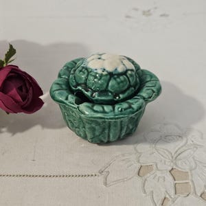 Può includere: Un portagioie in ceramica a forma di cavolo, con esterno verde e interno bianco e verde. La scatola è su una superficie bianca, con un bouquet di fiori rosso scuro sullo sfondo.