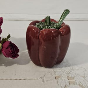 Könnte beinhalten: Ein glänzender, roter Keramikbehälter in Paprikaform mit einem grünen Stiel und Deckel. Der Behälter steht auf einer weißen Oberfläche, mit burgunderfarbenen Blumen im Hintergrund. Ein dekorativer Küchenartikel.