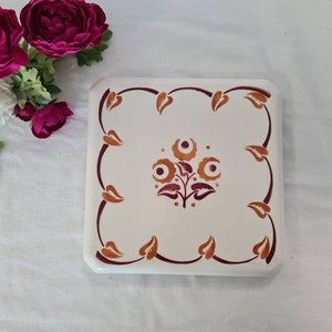 May include: A white ceramic square trivet with a burgundy and orange floral design. The trivet has a scalloped edge.