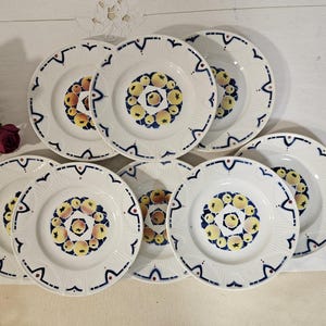 Może przedstawiać: Zestaw ośmiu białych ceramicznych talerzy z niebiesko-żółtym wzorem owoców. Każdy talerz ma centralny motyw owocowy otoczony niebieską obwódką z dekoracyjnymi elementami. Talerze są ułożone na białej powierzchni.