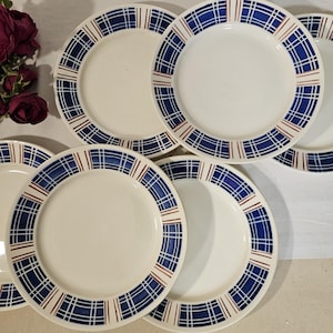 May include: A collection of white ceramic plates with a blue and red plaid pattern around the rims. The plates are arranged on a white surface, with a few red roses in the upper left corner.