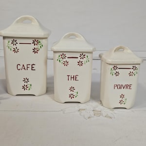 Op de afbeelding: Set van drie crèmekleurige keramische potten met deksels, met de opschriften "CAFÉ", "THÉ" en "POIVRE" in bordeauxrood. Elke pot heeft bordeauxrode bloemmotieven met groene bladeren. De deksels hebben een handvat.