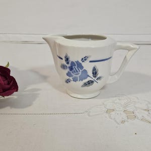 Peut inclure: Un pot à lait en céramique blanche orné d'un motif floral bleu représentant une rose unique avec des feuilles, accompagné d'une ligne bleue horizontale. Le pot possède un petit bec verseur et une anse angulaire, près de fleurs rose foncé.