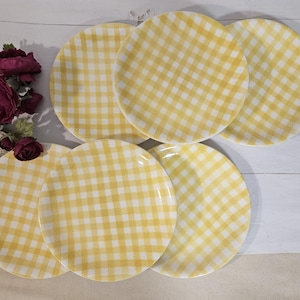 Peut inclure: Un ensemble de cinq assiettes rondes présentant un motif à carreaux vichy jaune et blanc, disposées en groupe. À côté des assiettes se trouve un petit bouquet de fleurs artificielles magenta foncé et blanches avec des feuilles et des tiges vertes.