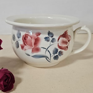 May include: White ceramic chamber pot with a handle and a floral rose design in shades of pink and blue. The roses are painted on the exterior of the pot, with a handle on the side.