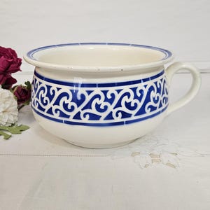 May include: A white ceramic chamber pot with a blue decorative pattern and a handle. The pot has a blue band around the rim and a matching design around the body. The pot is on a white surface.