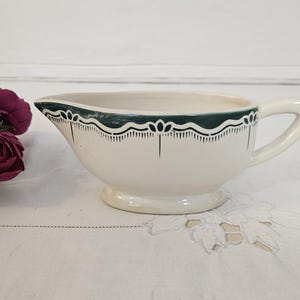 May include: Cream-coloured gravy boat with a dark green and black decorative border. The gravy boat has a handle and a small pedestal base. The rim is scalloped with a floral design. The gravy boat is on a white surface.