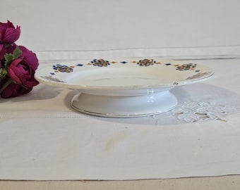May include: White ceramic cake stand with a raised base and a decorative rim. The plate features a repeating pattern of blue and gold floral designs. A bouquet of purple flowers is in the background.