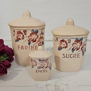 May include: Three beige ceramic kitchen canisters with lids, decorated with floral patterns in red, blue, and brown. The canisters are labelled "FARINE", "SUCRE", and "ÉPICES". The canisters are of varying sizes.