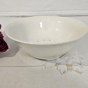 Puede incluir: Un cuenco de cerámica blanco con un borde ancho y pequeños orificios de drenaje. El cuenco está sobre una superficie blanca con un elemento floral decorativo y flores de color rojo oscuro.