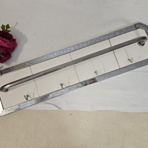 May include: A rectangular wall-mounted coat rack with a silver metal frame and a white tile background. It features a silver bar and three hooks for hanging items. The rack is approximately 24 inches long.