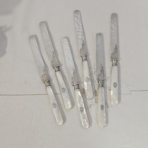Könnte beinhalten: Sechs antike Buttermesser mit Griffen aus Perlmutt und silberfarbenen Klingen. Jedes Messer hat eine dekorative silberfarbene Zwinge und ein Monogramm auf dem Griff. Die Messer sind auf einer weißen Oberfläche angeordnet.