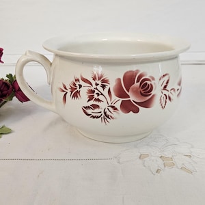 May include: A white ceramic chamber pot with a handle, decorated with burgundy floral designs. The pot has a wide rim and a rounded body. The background includes burgundy and white flowers.