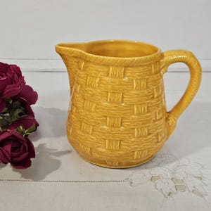 May include: A yellow ceramic pitcher with a woven basket design. The pitcher has a curved handle and a spout for pouring. A bouquet of dark red flowers and white flowers are on the left side of the pitcher.