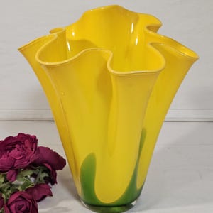 Könnte beinhalten: Eine leuchtend gelbe Glasvase mit gewelltem Rand und ausgestellter Form. Die Vase hat einen grünen Akzent am Boden und eine glänzende Oberfläche. Ein dekorativer Artikel für die Inneneinrichtung.