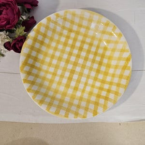 Peut inclure: Une assiette ronde en céramique ornée d'un motif à carreaux vichy jaune et blanc. L'assiette présente une finition lisse et brillante ainsi qu'un bord circulaire simple. Le motif se compose de lignes jaunes entrecroisées sur fond blanc.