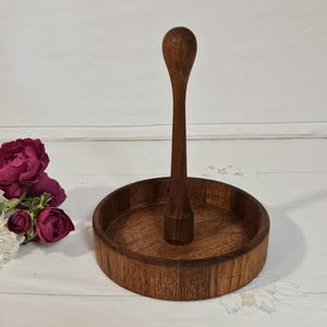 Puede incluir: Un portarrollos de papel de madera con base redonda y poste central alto. El soporte es de madera oscura y natural con un acabado liso. La base tiene aproximadamente 20 cm de diámetro.