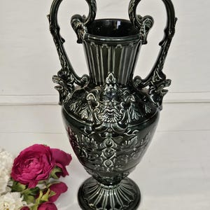 May include: A dark green ceramic vase with ornate handles and detailed relief carvings. The vase features a wide base, a bulbous midsection with a face design, and two curved handles. The vase is approximately 30 cm tall.