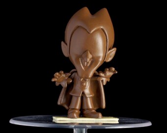 Peut inclure: Une figurine de vampire en chocolat brun avec une cape et des crocs, debout sur une base en plastique transparente.