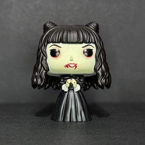 Puede incluir: Una figura de vinilo en blanco y negro de una mujer con cabello negro y un vestido negro. Tiene lápiz labial rojo y sangre en la boca.