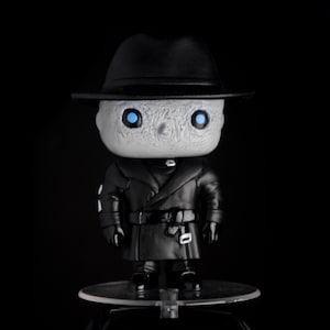 May include: A black and white vinyl figure of a character wearing a black coat and a fedora hat. The figure has blue eyes and a pale face.