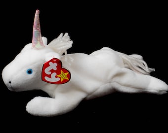 Könnte beinhalten: Ein weißes Plüsch-Einhorn-Spielzeug mit einem schimmernden, irisierenden Horn und blauen Augen. Das Einhorn hat eine weiße Mähne und einen weißen Schwanz sowie ein rotes herzförmiges Etikett mit gelbem Stern. Das Spielzeug liegt auf schwarzem Hintergrund.