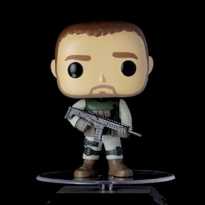 May include: A green and brown military-style figurine with a black gun, standing on a clear plastic base. The figurine has a large head with a brown beard and black eyes.