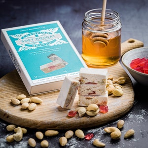May include: A box of soft nougat with cashew nuts, made with cashews, almonds, Turkish delight, and orange blossom honey. The box is blue and white with the words "Champion Soft Nougat with Cashew Nuts Reeves" printed on it. There are also some nougat pieces, cashews, and a jar of honey in the image.
