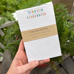 May include: A white notepad with horizontal lines and circular checkboxes. The top text reads every check off the list is a TINY WIN that deserves to be CELEBRATED. A brown paper band reads TINY WINS NOTEPAD 50 SHEETS - 4" x 6" - MADE IN USA.